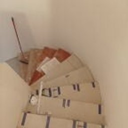 Wykończmy To - Schody w trakcie zabezpieczania przed pracami remontowymi, oklejone taśmą malarską, widok z góry na zakręcające biegi schodów. Częściowo widoczne płytki na stopniach. Ściany w kolorze złamanej bieli.