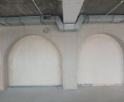 Surowe wnętrze z dwoma wnękami o łukowatym kształcie, betonowy sufit z widocznymi instalacjami wentylacyjnymi, ściany w jasnym, szarym odcieniu, na podłodze widoczne ślady po budowie.