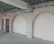 Wnętrze w trakcie prac wykończeniowych: surowy betonowy sufit, szare ściany z dwoma łukowatymi przejściami, widoczne przewody wentylacyjne pod sufitem, w tle fragment ściany z niedokończonymi...