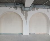 Dwa łukowate wnęki w ścianie, w trakcie prac wykończeniowych, widoczne ślady po gładzi szpachlowej, sufit z instalacją wentylacyjną, podłoga zabezpieczona folią.