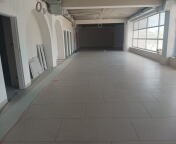 Pusta, jasna przestrzeń w trakcie prac wykończeniowych: surowy betonowy sufit, duże okna wzdłuż ściany, podłoga wyłożona płytkami, przy ścianie opierają się płyty gipsowo-kartonowe.