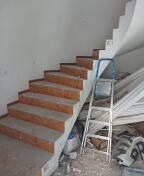 Betonowe schody z drewnianymi stopniami w trakcie montażu, widoczna drabina aluminiowa i płyty karton gips w tle, surowy stan wykończenia wnętrza.