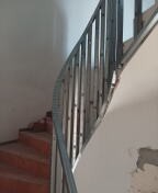 Klatka schodowa w trakcie remontu, widoczne schody z czerwonej terakoty i metalowa balustrada. Ściana częściowo przygotowana do dalszych prac wykończeniowych, widoczne fragmenty starej farby.