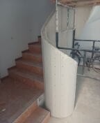 Klatka schodowa w trakcie budowy, widoczne schody z drewnianymi stopniami, obok kręta ściana z płyty kartonowo-gipsowej na metalowym stelażu, surowy stan wykończenia.