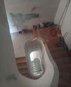 Klatka schodowa w trakcie gładzenia ścian, widoczne ślady po wcześniejszych pracach malarskich na górze ściany, surowe schody i prowizoryczne zabezpieczenia.