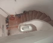 Wnętrze klatki schodowej w trakcie prac wykończeniowych, widok z góry. Widoczne drewniane schody kręcone, surowe ściany z widocznymi śladami po pracach budowlanych oraz narzędzia.