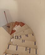 Schody w trakcie zabezpieczania przed pracami remontowymi, oklejone taśmą malarską, widok z góry na zakręcające biegi schodów. Częściowo widoczne płytki na stopniach. Ściany w kolorze złamanej bieli.