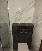 Obudowa stelaża podtynkowego WC wykończona czarnym marmurem z białymi żyłami, na tle białych płytek imitujących marmur. Widoczny fragment miski WC.