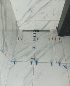 Układanie białych płytek imitujących marmur na ścianie, z użyciem poziomicy i klinów dystansowych w kolorze żółtym i niebieskim.