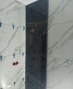 Fragment ściany z ułożonymi płytkami imitującymi marmur w kolorze białym i czarnym, widoczne niebieskie krzyżyki dystansowe i czerwone elementy instalacji, ujęcie z bliska.