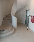 Wnętrze domu z krętymi schodami w trakcie prac wykończeniowych; ściany częściowo pokryte gładzią, widoczne narzędzia i niedokończone malowanie ścian w pobliżu wejścia.