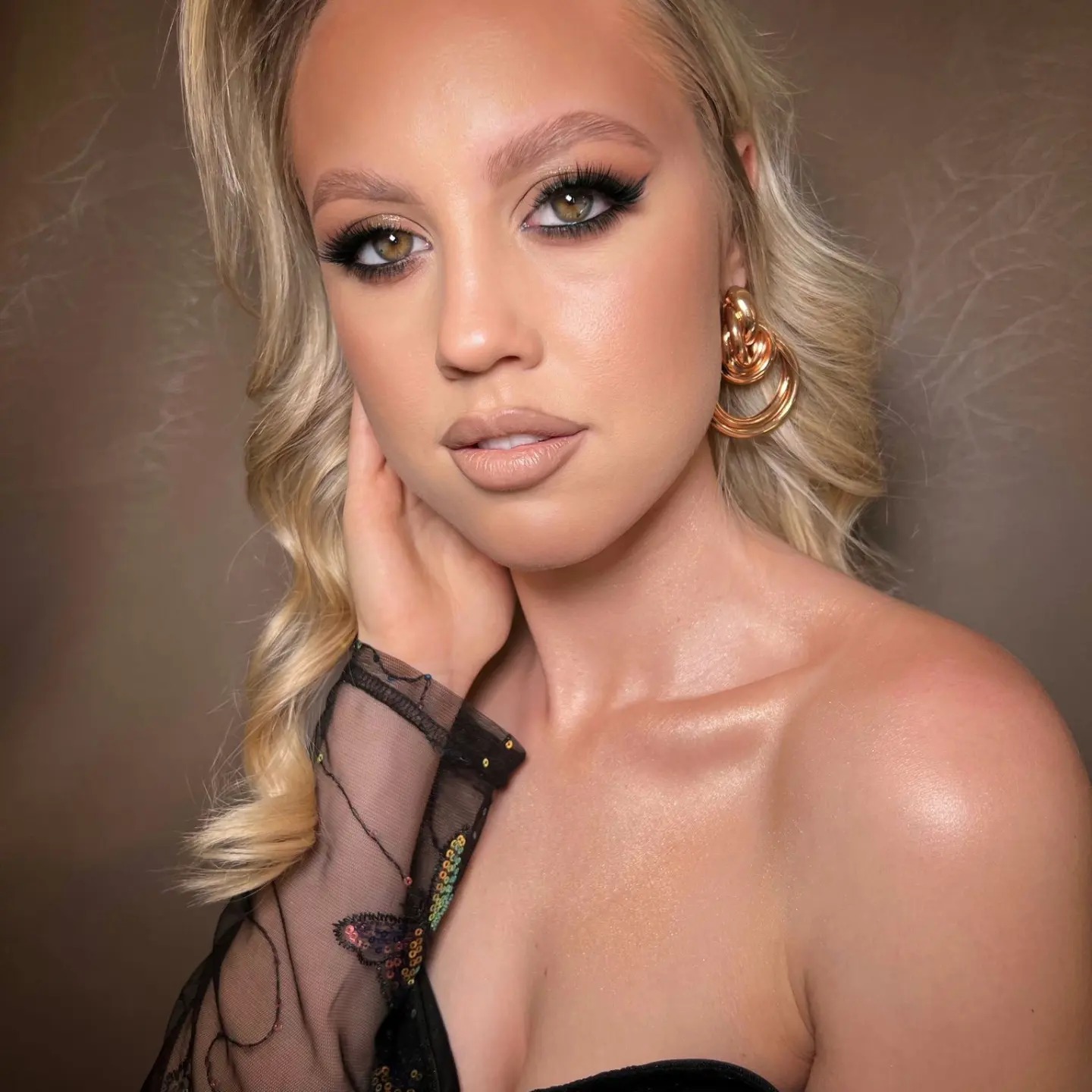 Portret blond modelki z profesjonalnym makijażem: wyraziste oko z eyelinerem, brwi podkreślone, usta w odcieniu nude, złote kolczyki, bluzka z siateczki z cekinami.