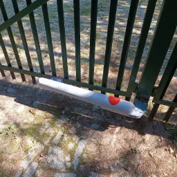 Zielona metalowa brama z zamontowanym szarym automatycznym napędem firmy Nice z pomarańczowym przyciskiem, widok z bliska na tle brukowanej powierzchni z zielenią.