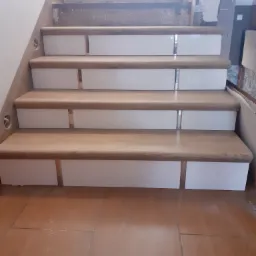 Wnętrze domu w trakcie remontu: widok na drewniane schody z białymi podstopnicami, częściowo wykończone, z widocznymi śladami prac budowlanych na g&oacute;rnych stopniach i podłodze.