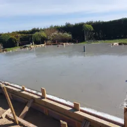 Świeżo wylany, gładki fundament z szalunkiem drewnianym i wystającym pionowym rurociągiem kanalizacyjnym, widok na otaczający ogród z zielonymi krzewami i drzewami w tle.