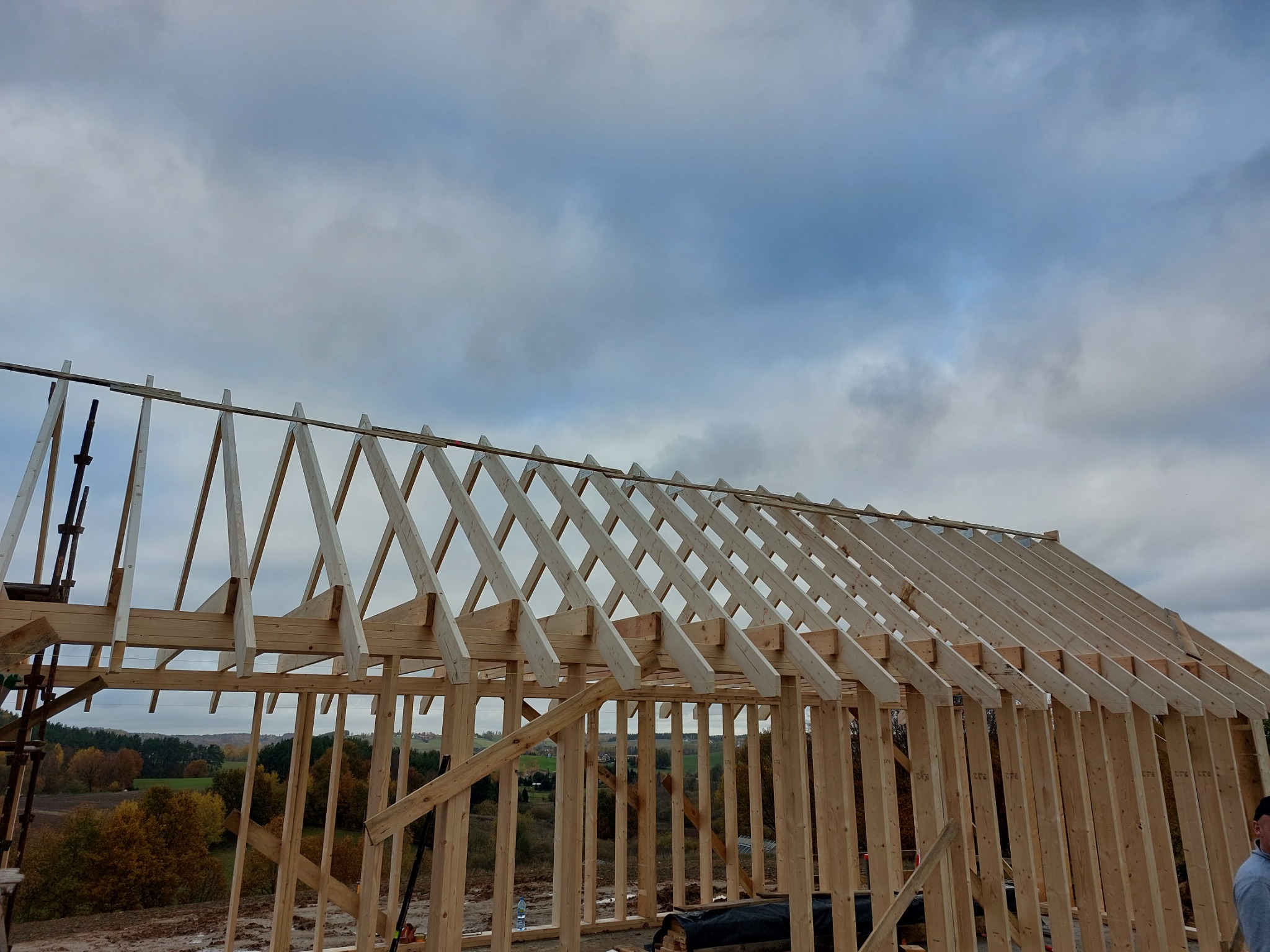 Konstrukcja drewniana dachu w trakcie budowy domu, widok z dołu na tle pochmurnego nieba i jesiennego krajobrazu w oddali.