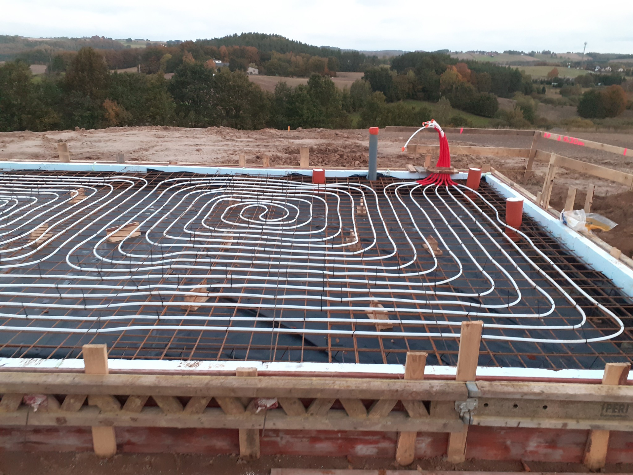Przygotowanie płyty fundamentowej z instalacją ogrzewania podłogowego, widoczne białe rurki ułożone w spiralę na zbrojeniu, izolacja termiczna po bokach i drewniane szalunki, w tle krajobraz wiejski.