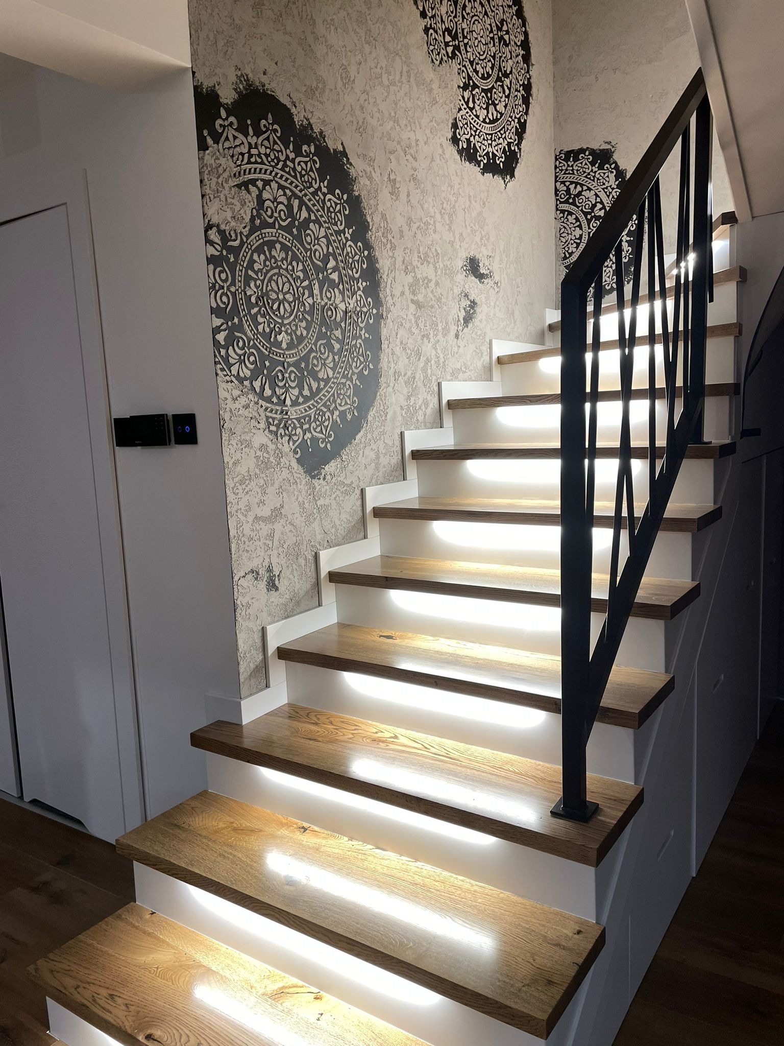 Nowoczesne schody drewniane z podświetleniem LED, czarna balustrada, ściana z dekoracyjnym wzorem mandali.