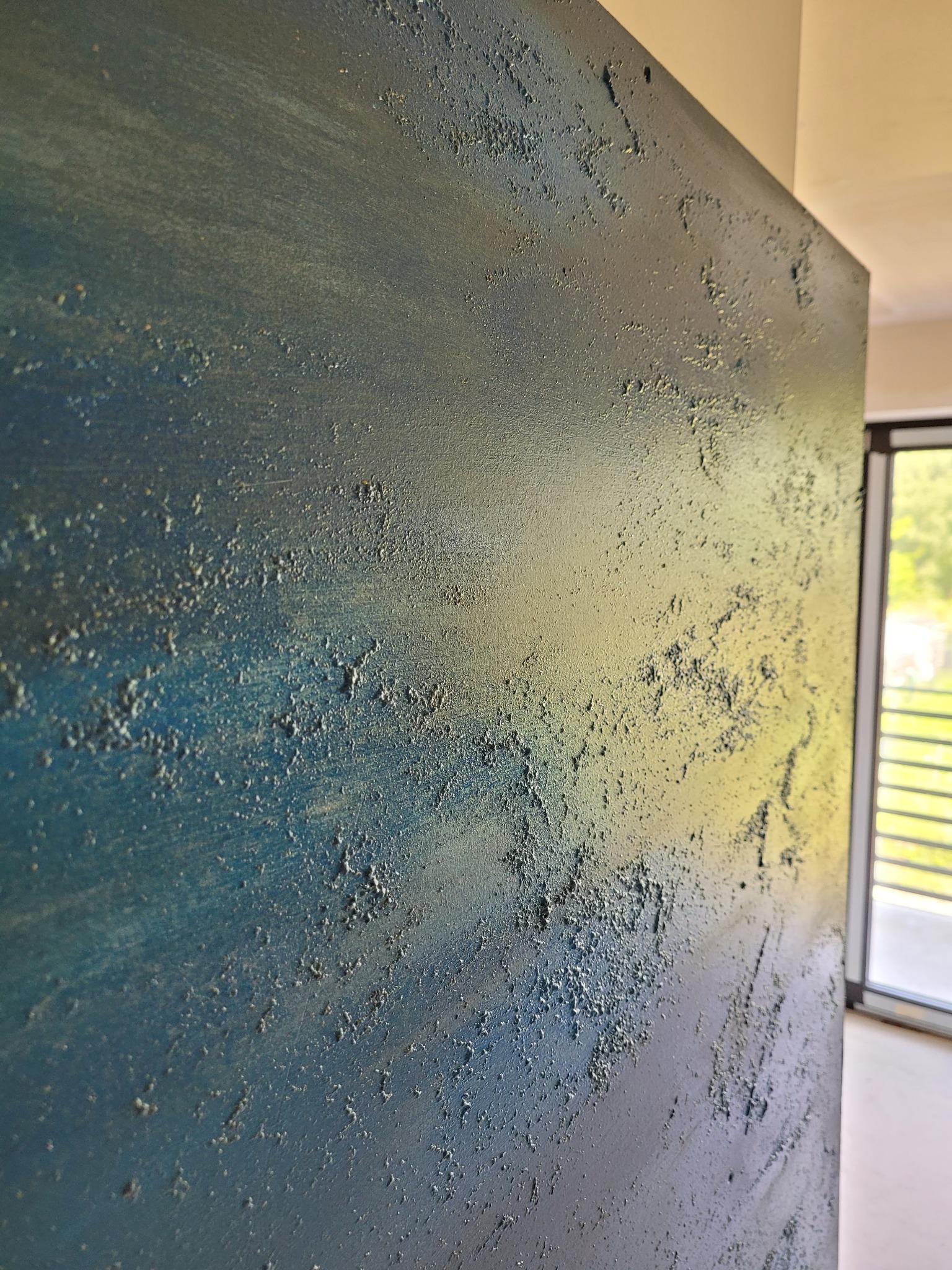Niebiesko-szara ściana z wyraźną, nieregularną fakturą, przypominającą efekt dekoracyjny z widocznymi nierównościami i drobnymi grudkami, oświetlona z prawej strony, z widocznym fragmentem okna...