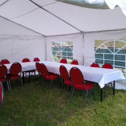 Wnętrze białego namiotu z rozstawionymi stołami przykrytymi białymi obrusami i czerwonymi krzesłami, widok przez okno namiotu na zewnątrz.