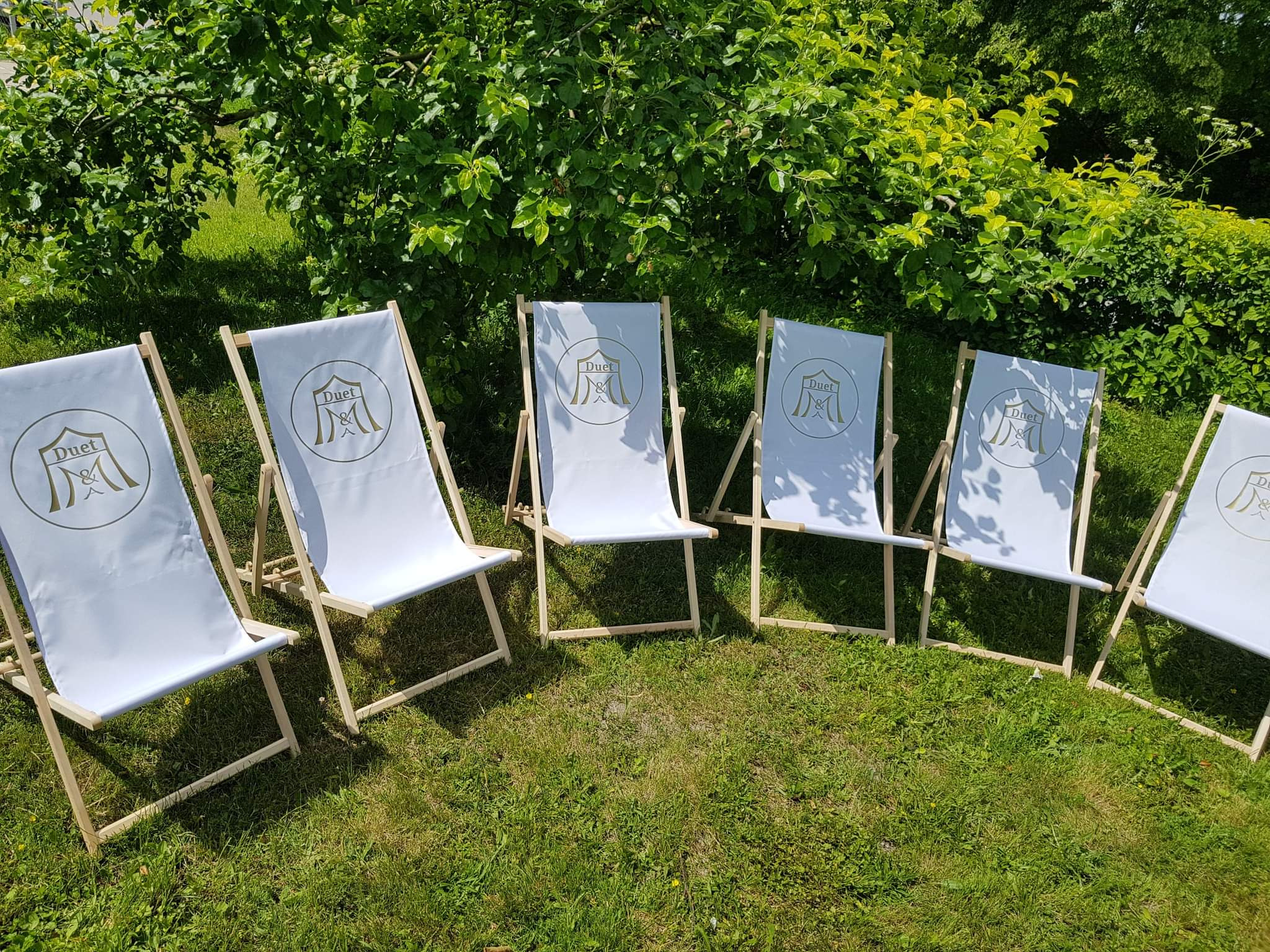 Rząd białych, drewnianych leżaków z nadrukowanym logo 'Duet' na zielonej trawie, w tle krzewy.