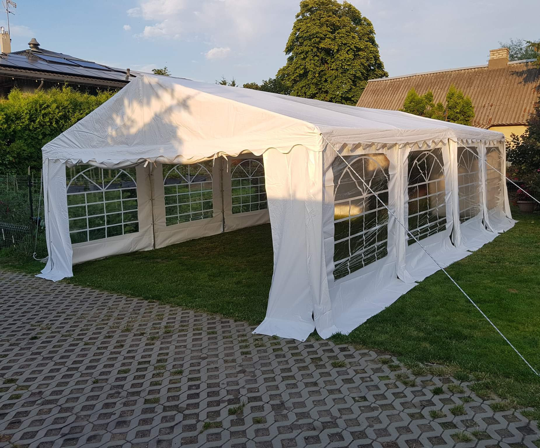 Rozłożony biały namiot imprezowy z oknami, ustawiony na trawie obok betonowej kostki, w tle domy i drzewa.