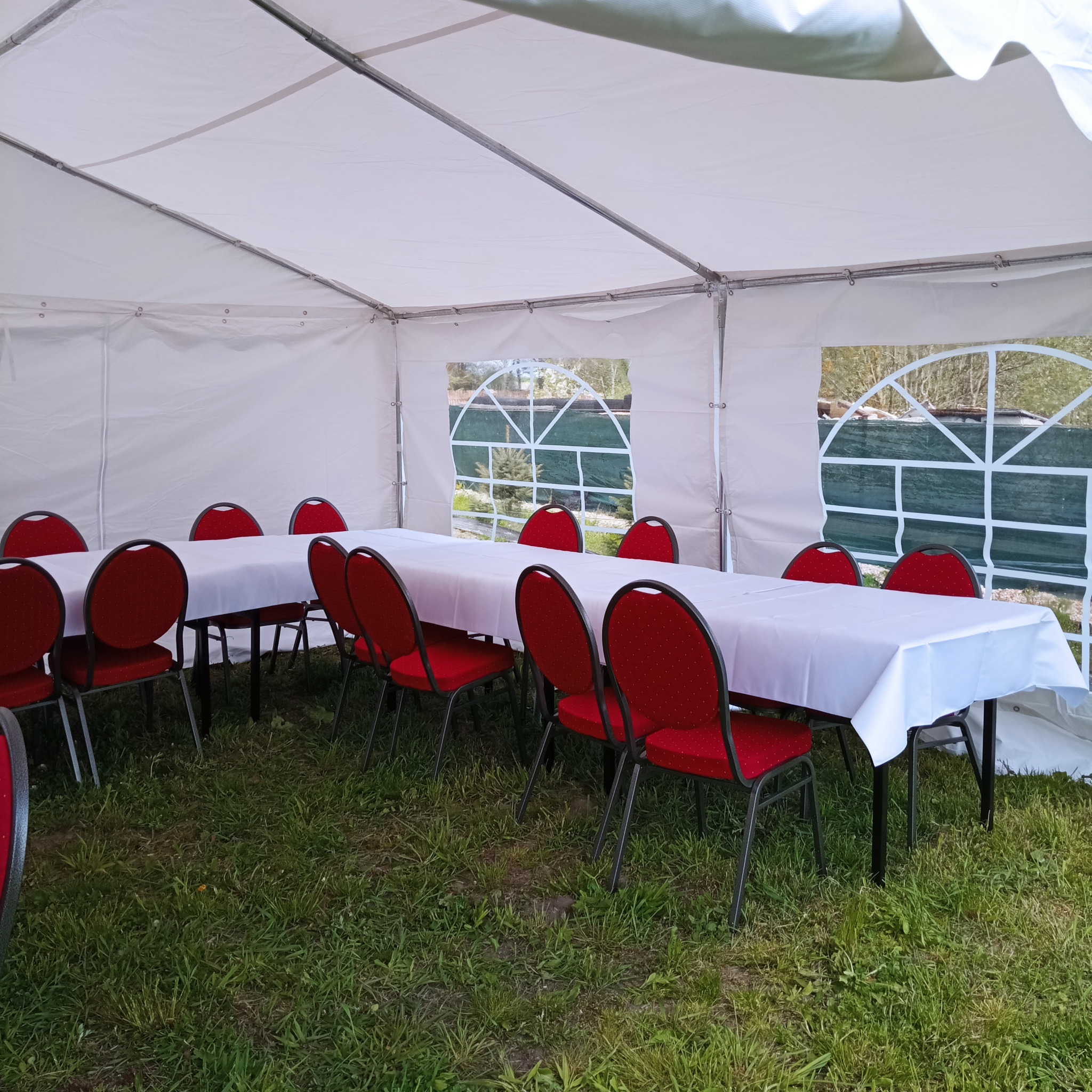 Wnętrze białego namiotu z rozstawionymi stołami przykrytymi białymi obrusami i czerwonymi krzesłami, widok przez okno namiotu na zewnątrz.