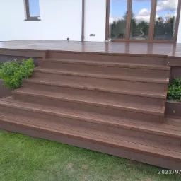 Brązowe schody z desek prowadzące na taras przy białym domu, widoczne rośliny i odbicie chmur w oknach.