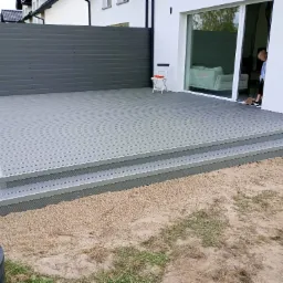 Nowoczesny taras z szarych desek kompozytowych z trzema stopniami, ogrodzony szarą ścianką, z białym krzesłem składanym i osobą zamiatającą podłogę przy wejściu do domu.