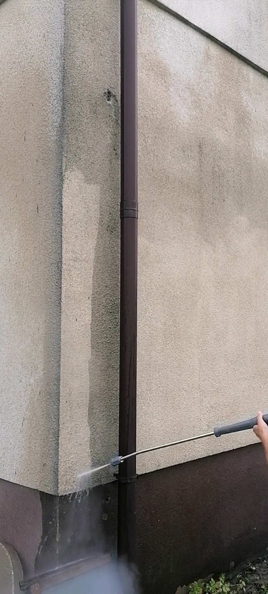 Mycie elewacji budynku z brązową rynną, widoczny efekt czyszczenia strumieniem wody pod ciśnieniem, fragment elewacji już oczyszczony, a na dole widoczne zabrudzenia.