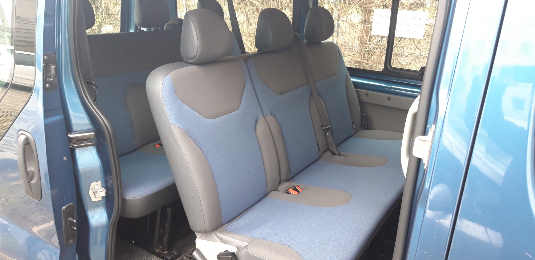 Wnętrze niebieskiego busa z widocznymi dwoma rzędami foteli w odcieniach szarości i błękitu, z otwartymi drzwiami bocznymi.