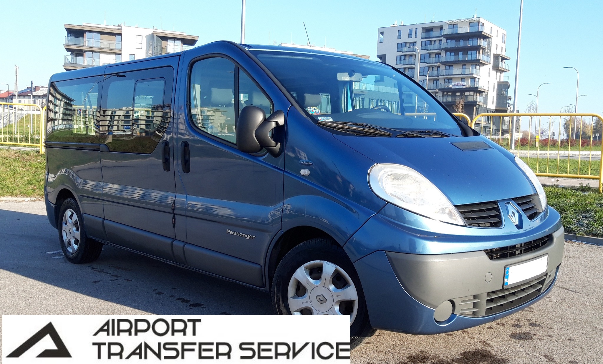 Niebieski van Renault Trafic Passenger z napisem 'Passenger' na błotniku, zaparkowany na parkingu z widokiem na nowoczesne apartamentowce w tle.