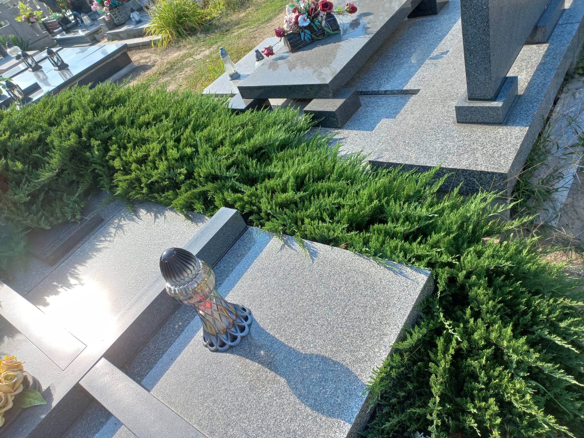 Granitowy nagrobek z dekoracyjną, niską zielenią iglastą, lampionem i kwiatami, na cmentarzu w słoneczny dzień.