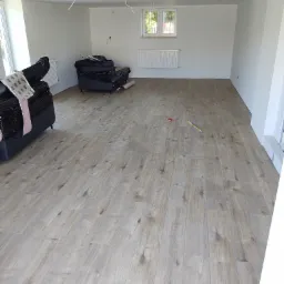 Jasne panele podłogowe ułożone w nowym salonie, meble zabezpieczone folią, widoczne narzędzia pomiarowe.