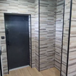 Wnętrze przedpokoju z dekoracyjną, poziomą okładziną ścienną w odcieniach beżu i szarości, oraz podłogą imitującą drewno, z czarną, metalową ościeżnicą drzwi wejściowych.