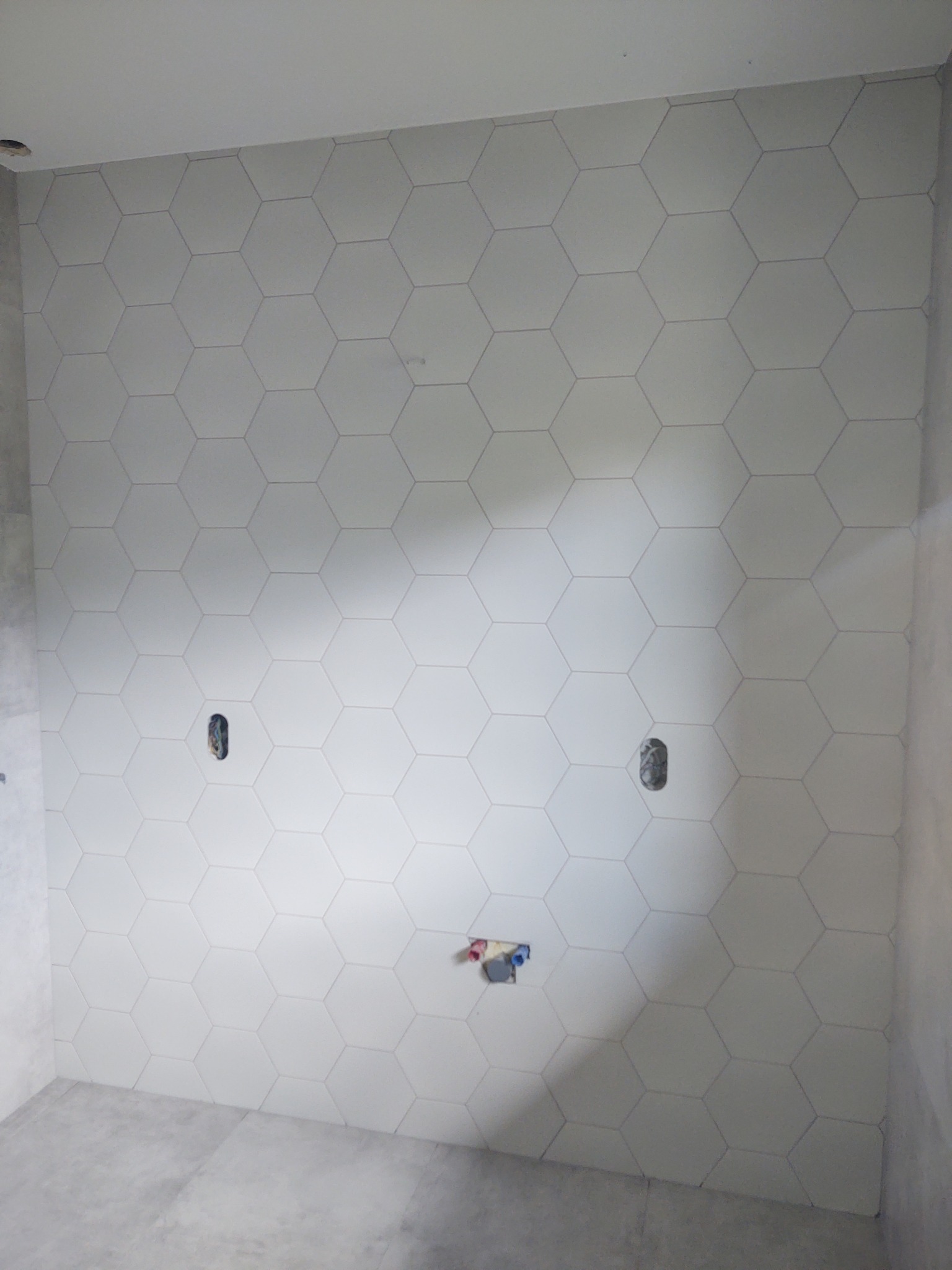 Ściana łazienki wyłożona białymi, heksagonalnymi płytkami ceramicznymi z widocznymi otworami na gniazdka elektryczne i przyłącza wodne, ukazująca etap prac wykończeniowych.