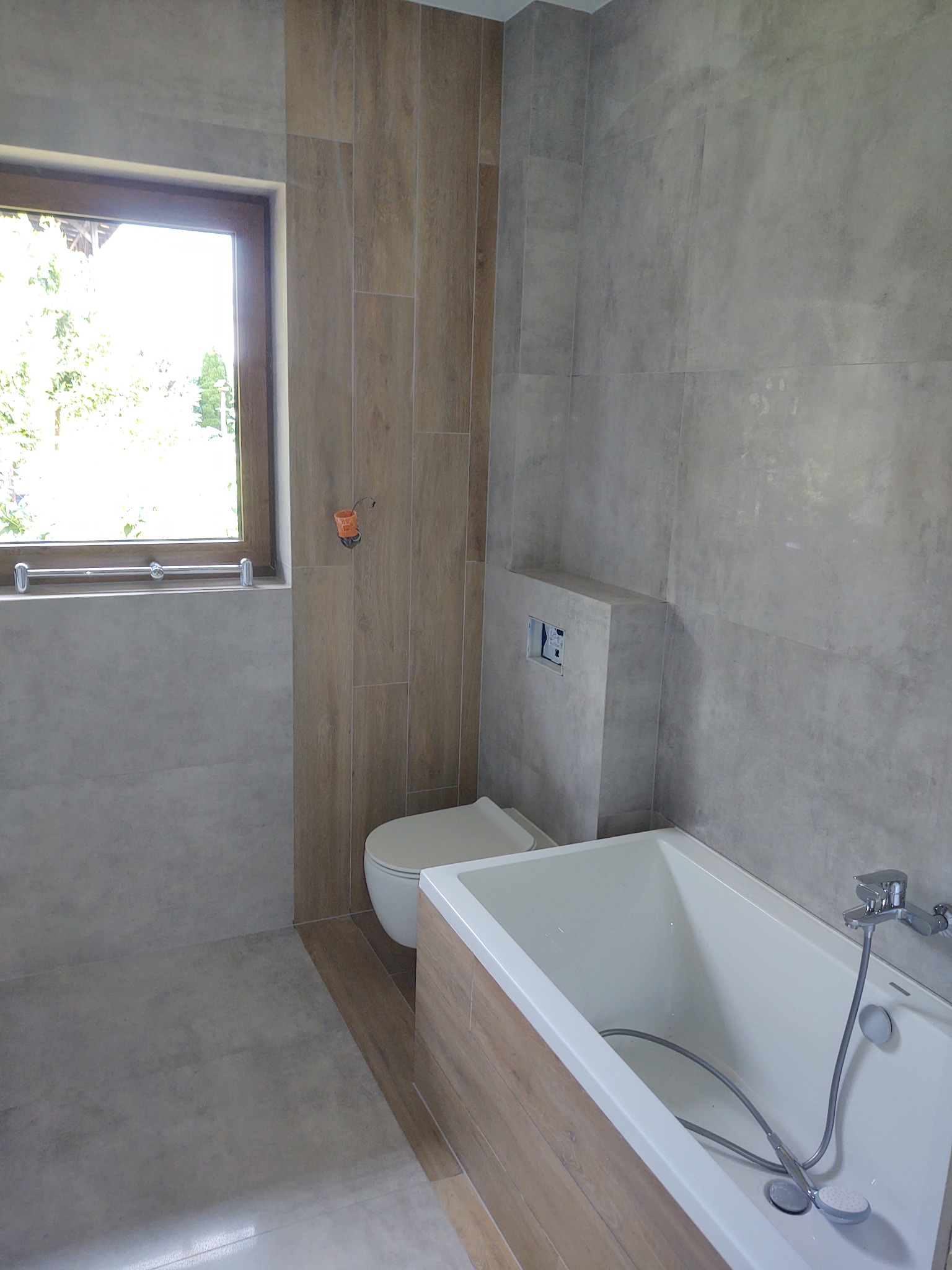 Nowoczesna łazienka z wanną obudowaną panelami imitującymi drewno, szare płytki na ścianach i podłodze, oraz wisząca toaleta z podtynkowym systemem spłukiwania w trakcie montażu.