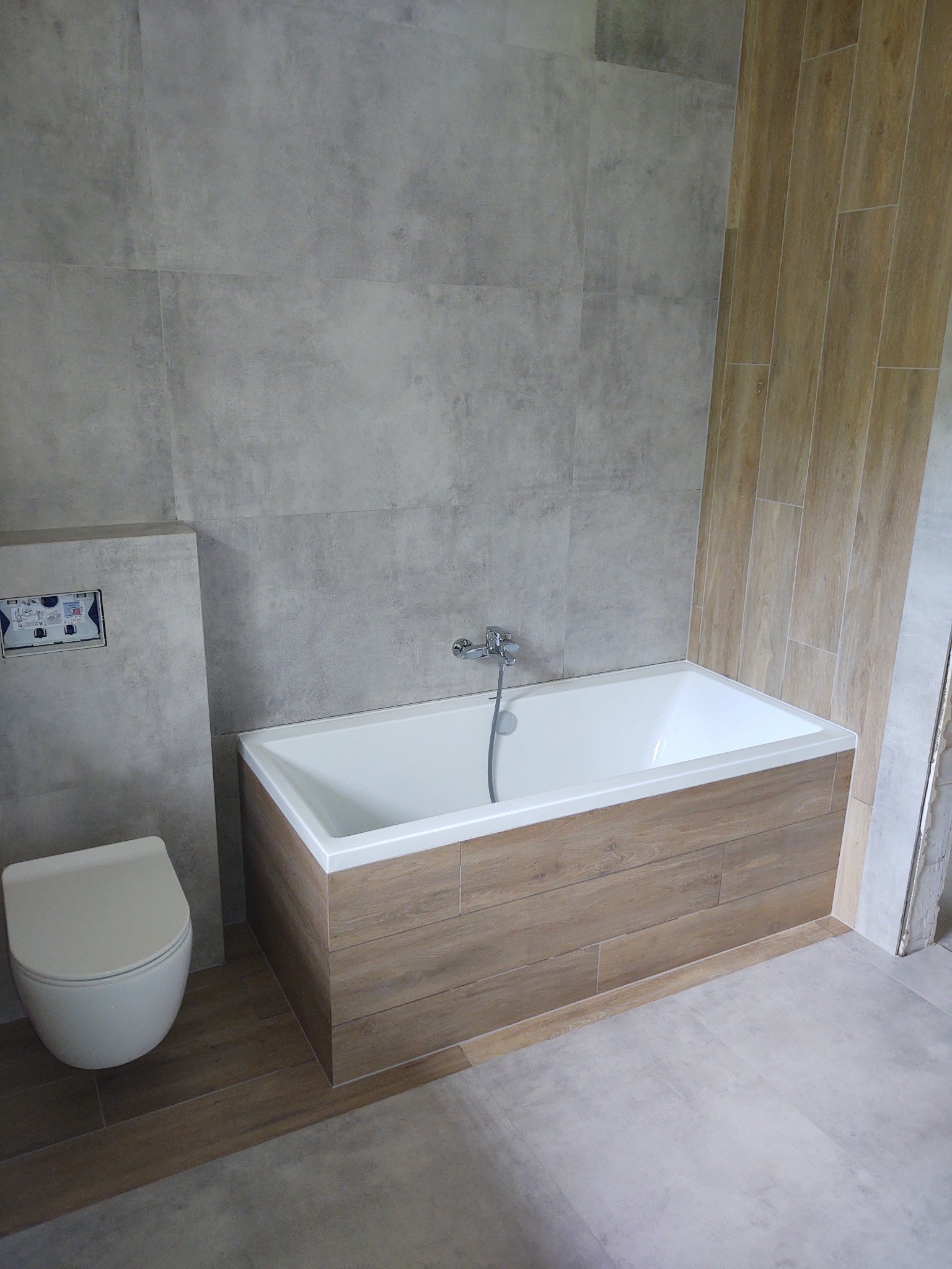 Nowoczesna łazienka z wanną obudowaną płytkami imitującymi drewno, szare płytki ścienne i podłogowe oraz podwieszana toaleta.
