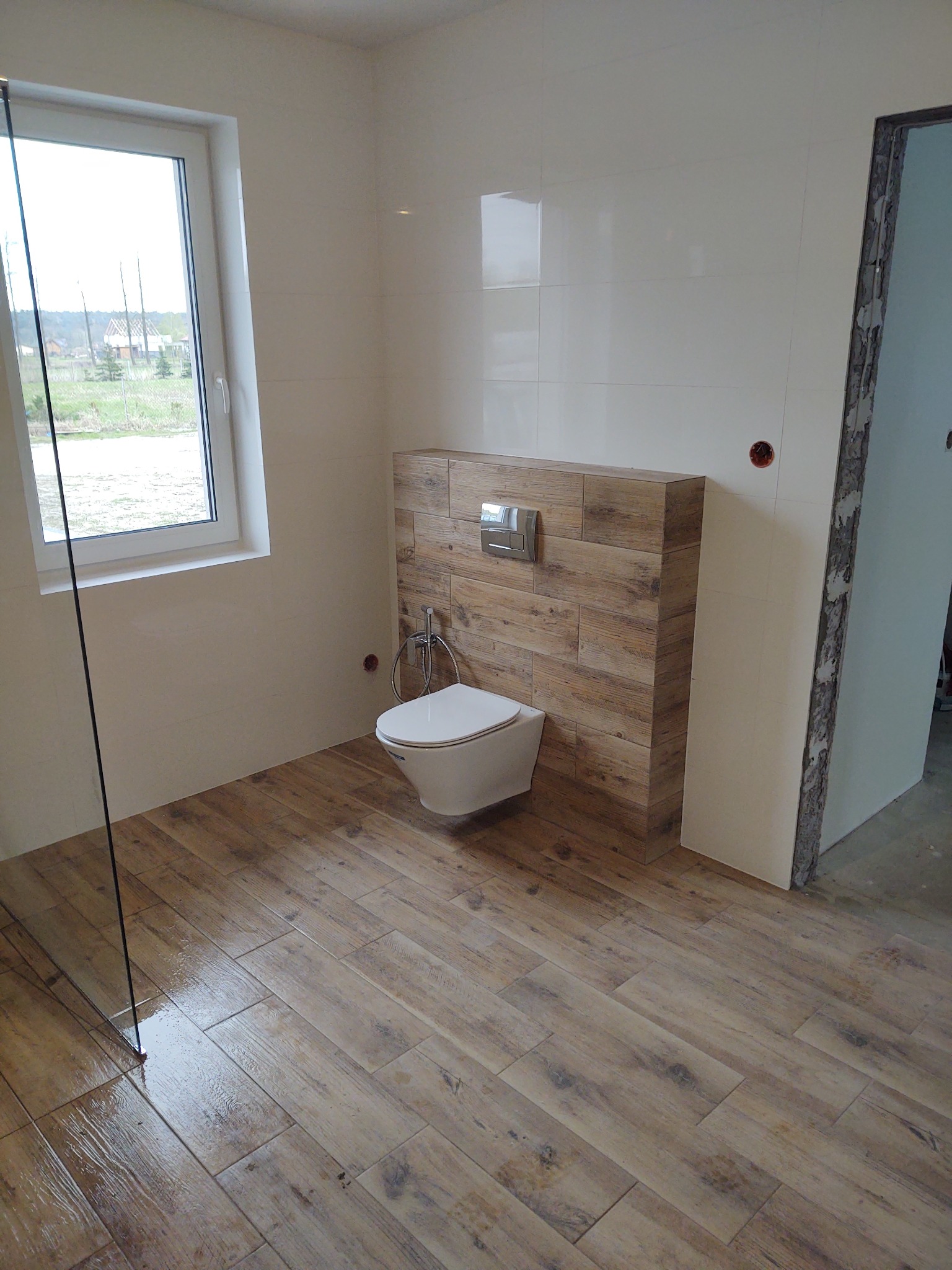 Nowoczesna łazienka z białymi płytkami ściennymi, drewnopodobną zabudową toalety i podłogą z płytek imitujących drewno, widok na okno i fragment niedokończonego wejścia.