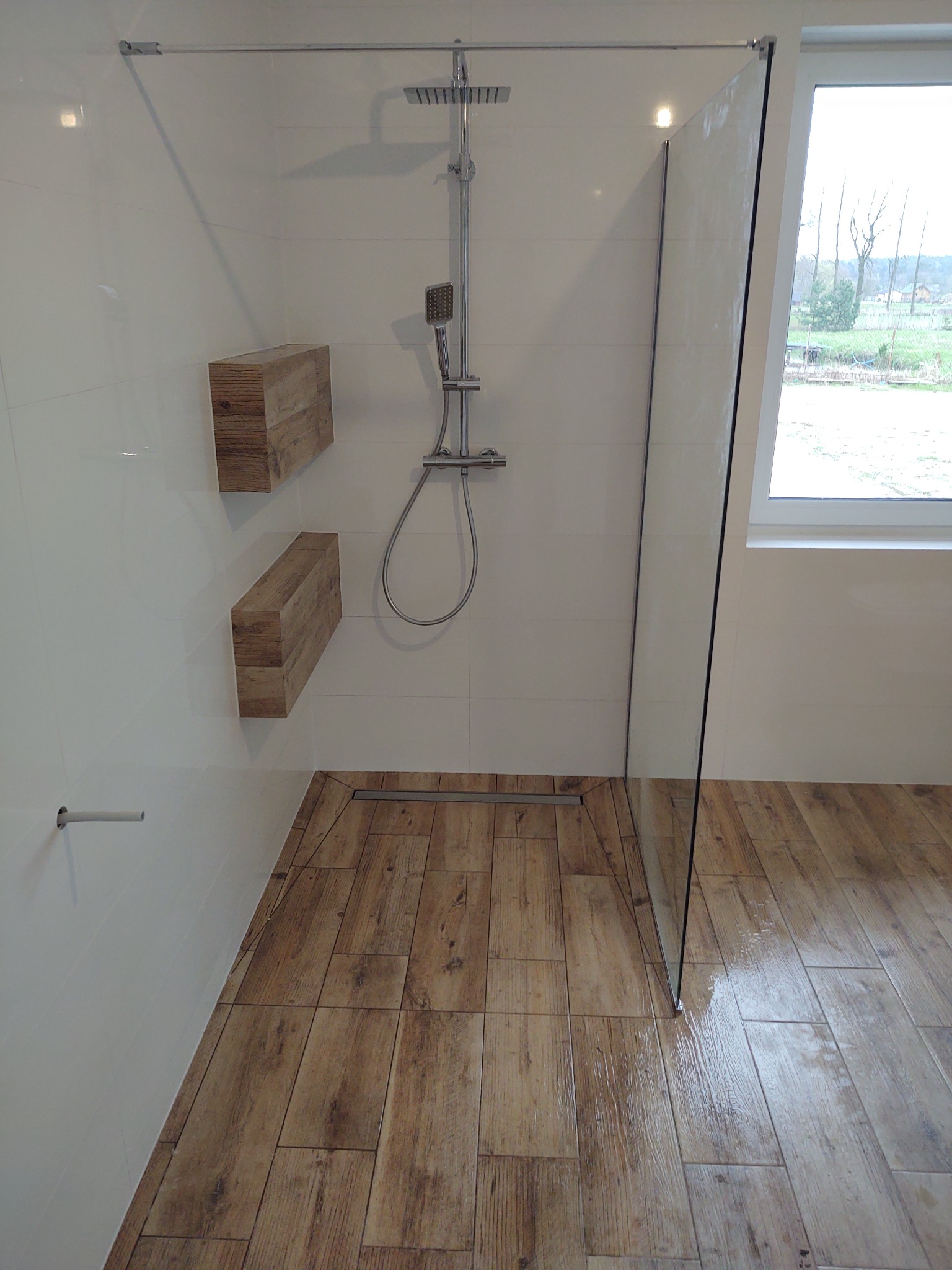 Nowoczesna kabina prysznicowa typu walk-in z drewnopodobną podłogą i półkami z surowego drewna, widok na zewnątrz przez okno.