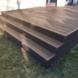 Nowoczesne, brązowe schody z desek kompozytowych prowadzące na taras, widoczne na tle trawnika i elewacji budynku.