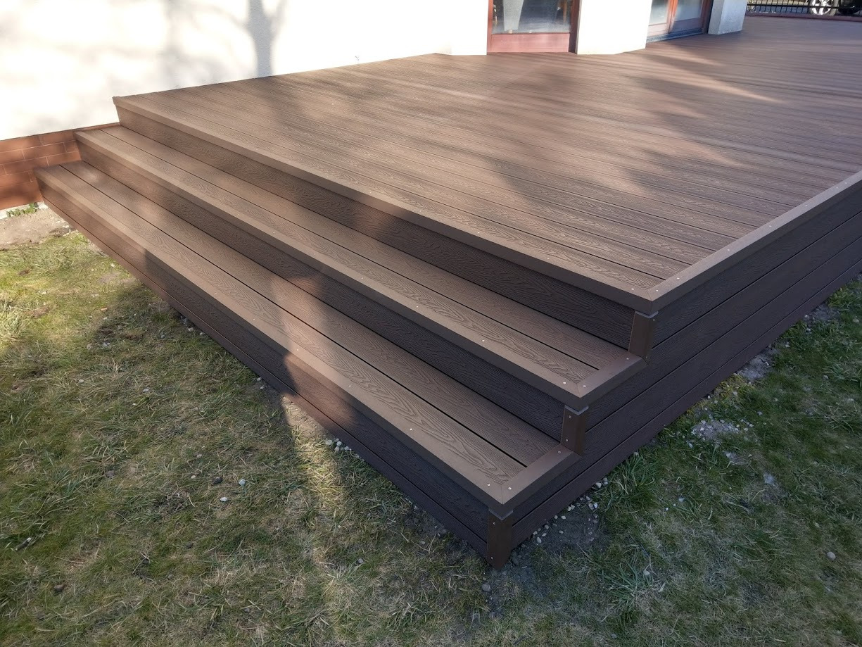 Nowoczesne, brązowe schody z desek kompozytowych prowadzące na taras, widoczne na tle trawnika i elewacji budynku.