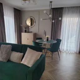 Salon z zieloną sofą, okrągłym stołem z marmurowym blatem i złotą podstawą, oraz szafką z lustrem w tle, oświetlony nowoczesnymi lampami.