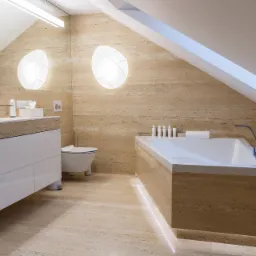 Jasna łazienka na poddaszu z beżowymi płytkami trawertynowymi, podświetlana wanna, biała szafka pod umywalką i okrągłe lampy ścienne.