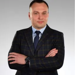 Portret mężczyzny w ciemnej, kraciastej marynarce, jasnej koszuli i granatowym krawacie, z zegarkiem na ręku i obrączką na palcu, na szarym tle.