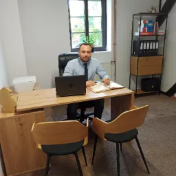 Mężczyzna w jasnej koszuli i krawacie siedzi za biurkiem w jasnym biurze, pisząc w otwartym notatniku obok laptopa; za nim okno i regał z dokumentami.