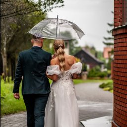 kierophoto - Młoda para w deszczu idzie ścieżką pod przezroczystym parasolem. Panna młoda w białej sukni z odkrytymi plecami, pan młody w ciemnozielonym garniturze.