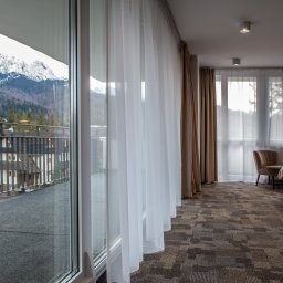 kierophoto - Jasny pokój hotelowy z balkonem i widokiem na góry. Przez okno widać ośnieżone szczyty i zabudowę. Wnętrze z beżowymi zasłonami i dywanem w geometryczne wzory.