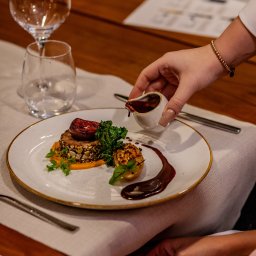 kierophoto - Elegancko podane danie z figą i sosem na białym talerzu, z ręką polewającą sosem z dzbanka. W tle kieliszki i drewniany stół. Fine dining.