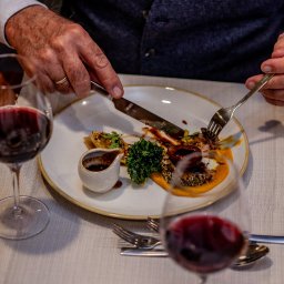 kierophoto - Elegancko podane danie na talerzu, krojone nożem i widelcem przez mężczyznę w kamizelce, z kieliszkami czerwonego wina w tle. Fine dining w restauracji.